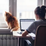 Kvinde sidder ved bærbar computer foran et vindue, hvor en kat sidder og kigger ud